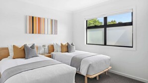 A modern hotel room with two single beds, a large window with blinds, a wall-mounted artwork, and a bedside table with a lamp. - St Helens I Stunning Family Townhouse in Geelong (Rippleside)
