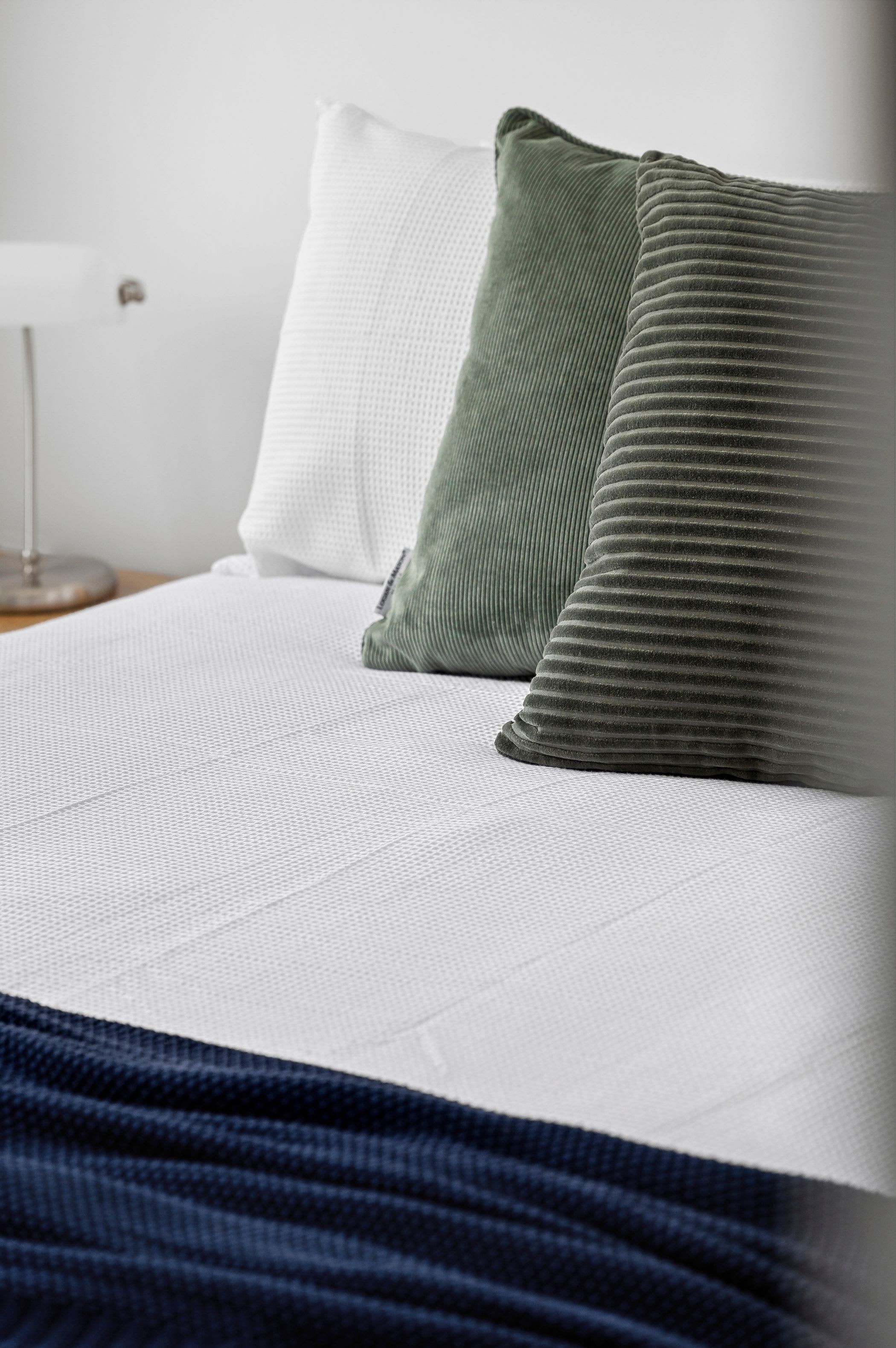 Un lit avec des oreillers blancs et verts, une couvre-lit blanche à motifs et une couverture bleu foncé.