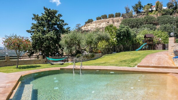 A clear swimming pool with a stone deck, a hammock, and a playground with a slide.