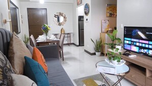 A modern living room with a sofa, a dining table with chairs, a TV, and decorative plants. - Deluxe 1BR 27th floor in Taguig/BGC w/balcony, amazing view of bay, wi-fi+55" TV (Taguig)