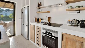 A modern kitchen with a refrigerator, oven, and wooden cabinets. There are wooden shelves above the counter holding various kitchen items. - Jujube - Javea, Costa Blanca (Javea)