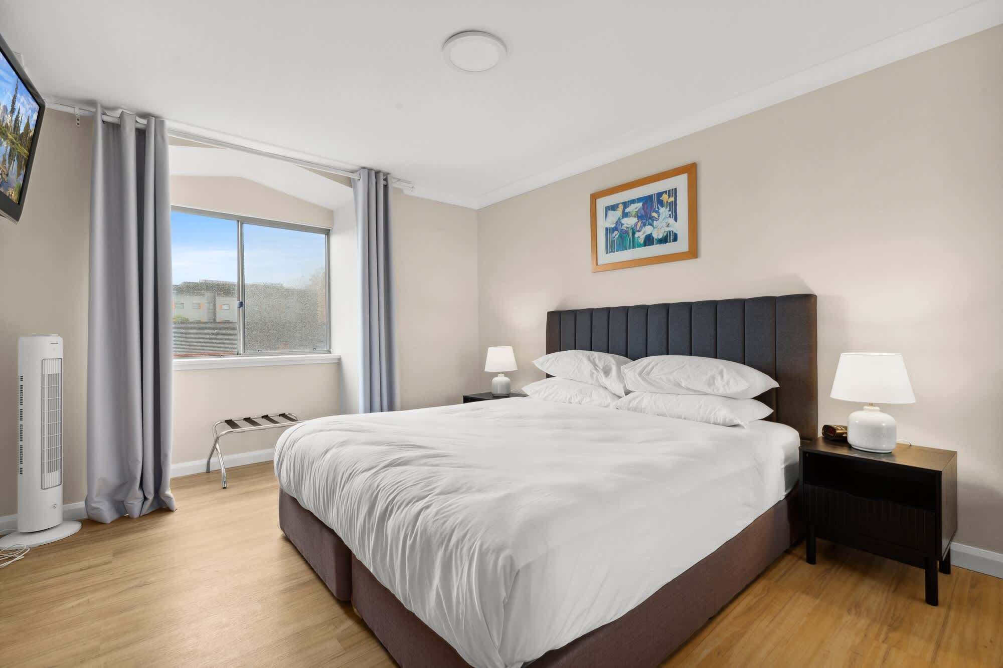 A hotel room with a large bed, two bedside tables with lamps, a window with curtains, a radiator, and a painting on the wall.