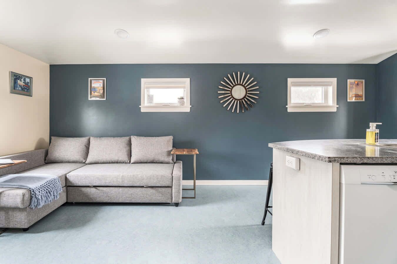 A modern living room with a grey sectional sofa, a small wooden side table, and a kitchen area with a countertop and dishwasher.