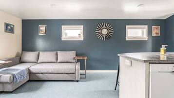 A modern living room with a grey sectional sofa, a small wooden side table, and a kitchen area with a countertop and dishwasher.
