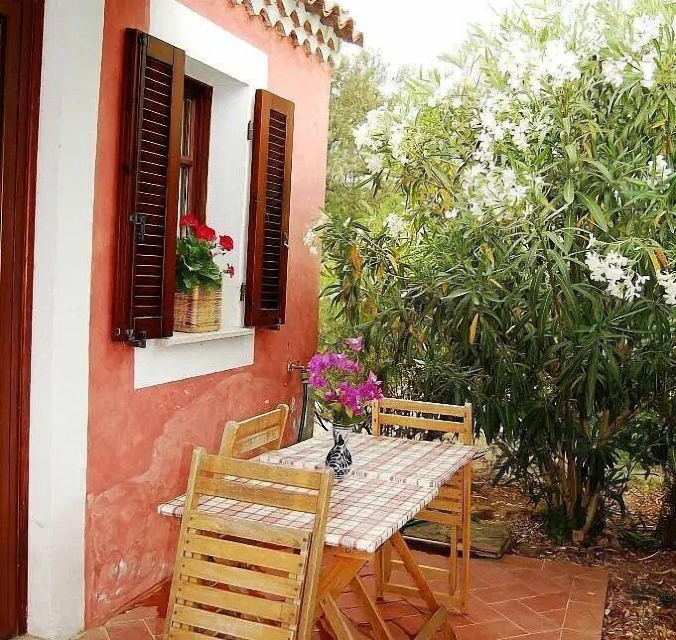 A wooden table and chairs set outside a building with a window displaying red flowers.