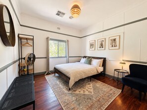 A bedroom with a bed, a chair, a desk, a standing fan, a window with curtains, and framed pictures on the wall. - 4BR Central Dubbo Family Home, 2 Living Areas, Large Backyard, Sleeps 8 (Dubbo)