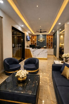 A hotel lobby with a reception desk, two armchairs, a sofa, a coffee table, and a ceiling fan. - Kennedia International Hotel And Suites (Varanasi)