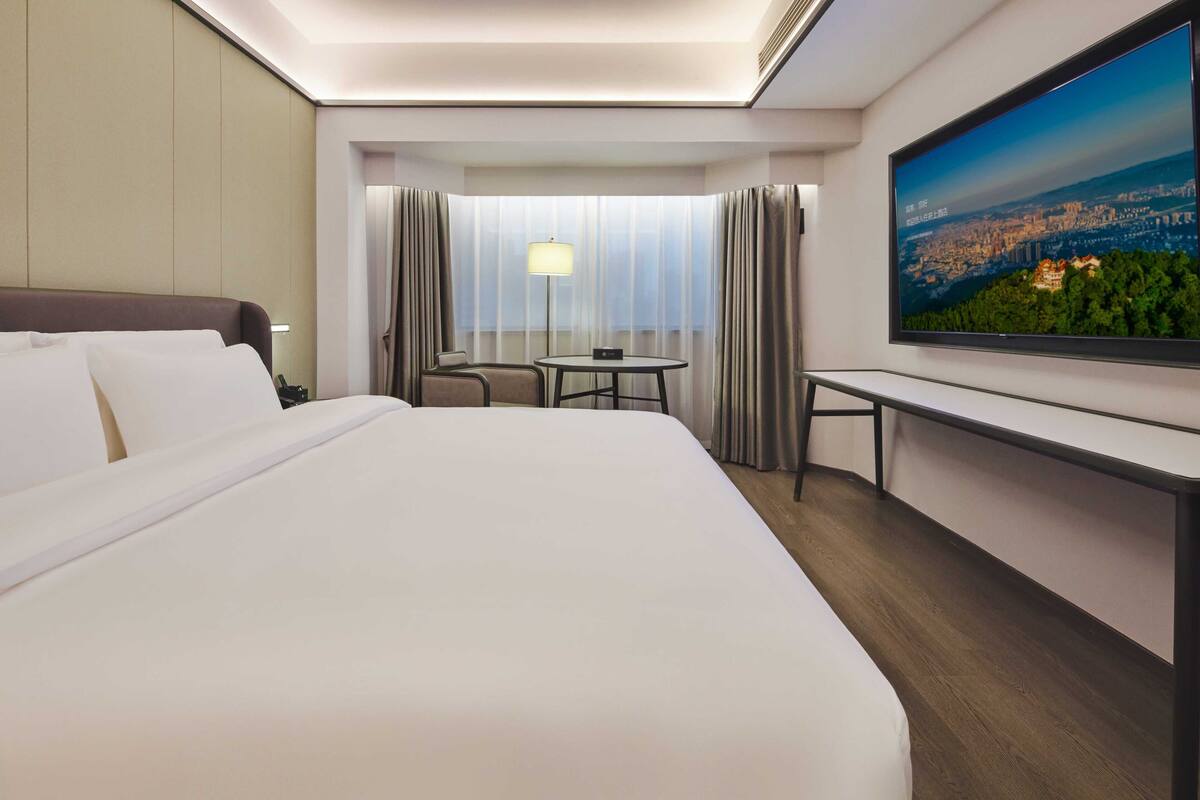 A hotel room with a large bed, a desk, a chair, a TV, and a window with curtains.