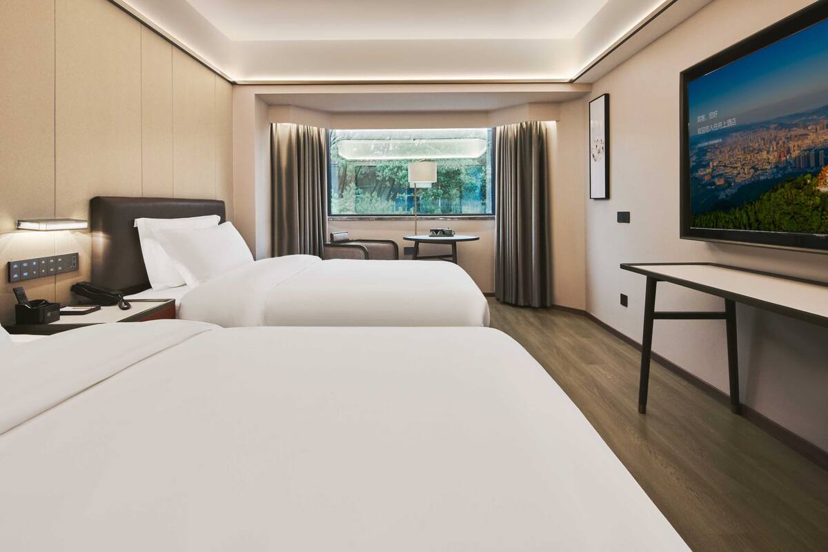 A hotel room with two beds, a desk, a chair, a TV, and a window with curtains.