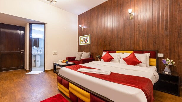 A hotel room with a wooden paneled wall, a bed with red and white bedding, a red sofa, a bedside table with a vase of flowers, and a bathroom visible through an open door. - Nook Inn & Suites (Gurugram)