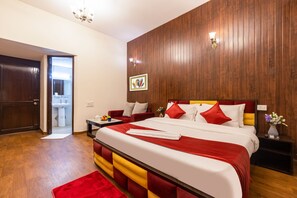 A hotel room with a wooden paneled wall, a bed with red and white bedding, a red sofa, a bedside table with a vase of flowers, and a bathroom visible through an open door.