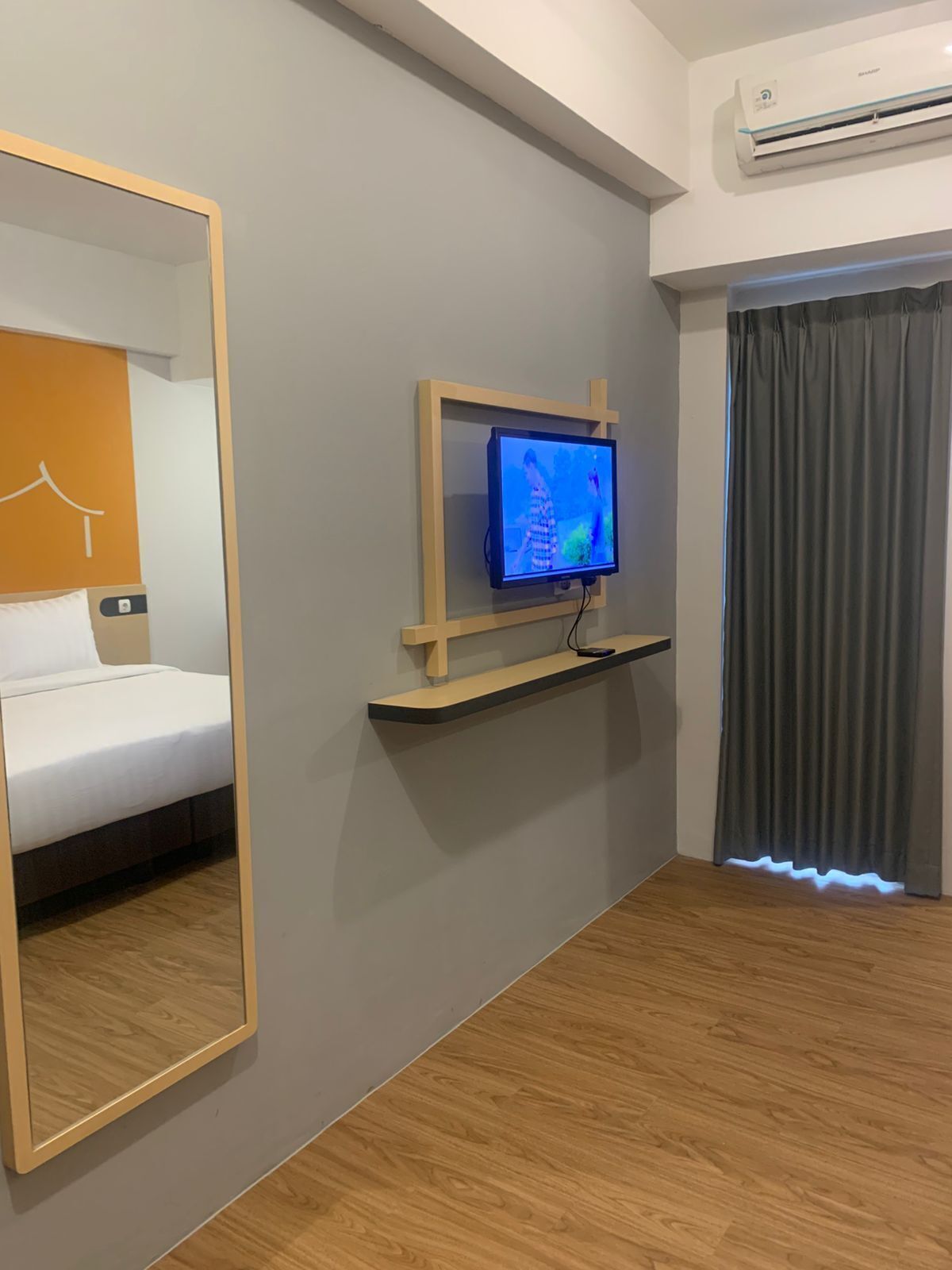 A hotel room with a bed, a TV mounted on a wooden shelf, a large mirror, and an air conditioning unit.