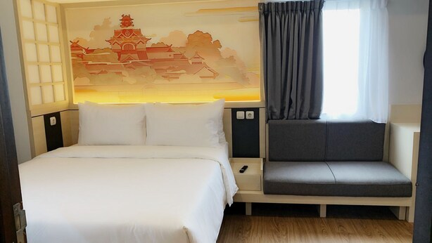 A hotel room with a bed, a sofa, a wall-mounted TV, a window with curtains, and a decorative wall mural. - Nemuru Stay BTC (Bekasi)