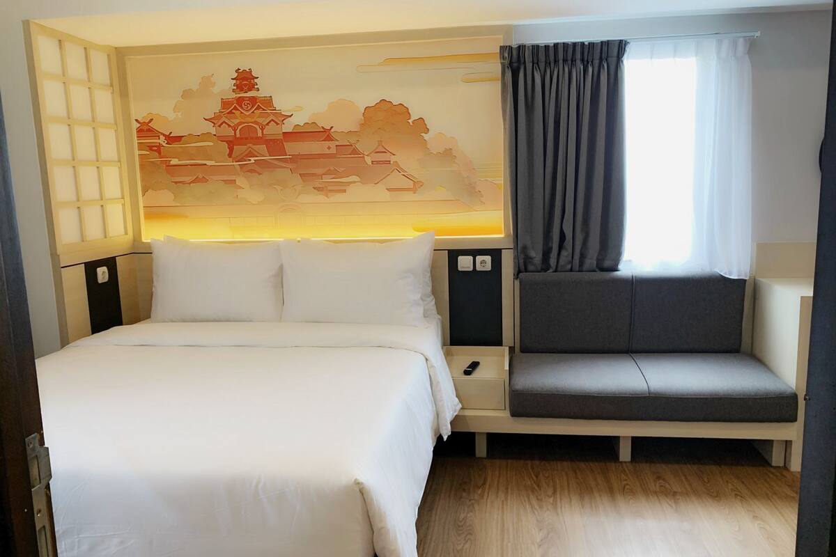 A hotel room with a bed, a sofa, a wall-mounted TV, a window with curtains, and a decorative wall mural.