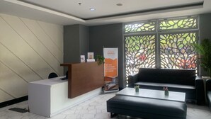 A reception area with a wooden desk, a white reception counter, a black sofa, and a decorative window with a tree pattern. - Nemuru Stay BTC (Bekasi)