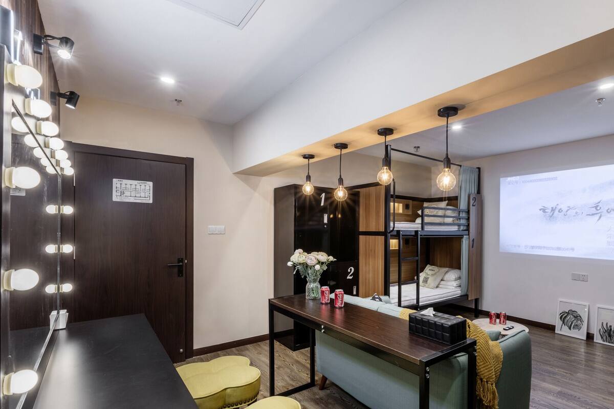 Habitación de hotel con espejo de tocador, una mesita con un florero y una cama litera, además de una pantalla de proyector mostrando texto.