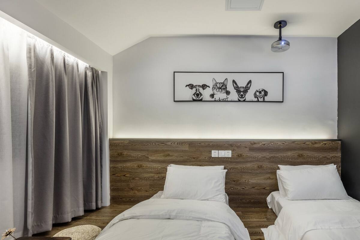 Un dormitorio moderno con dos camas, un cabecero de madera, una lámpara en el techo y una obra de arte enmarcada sobre las camas.