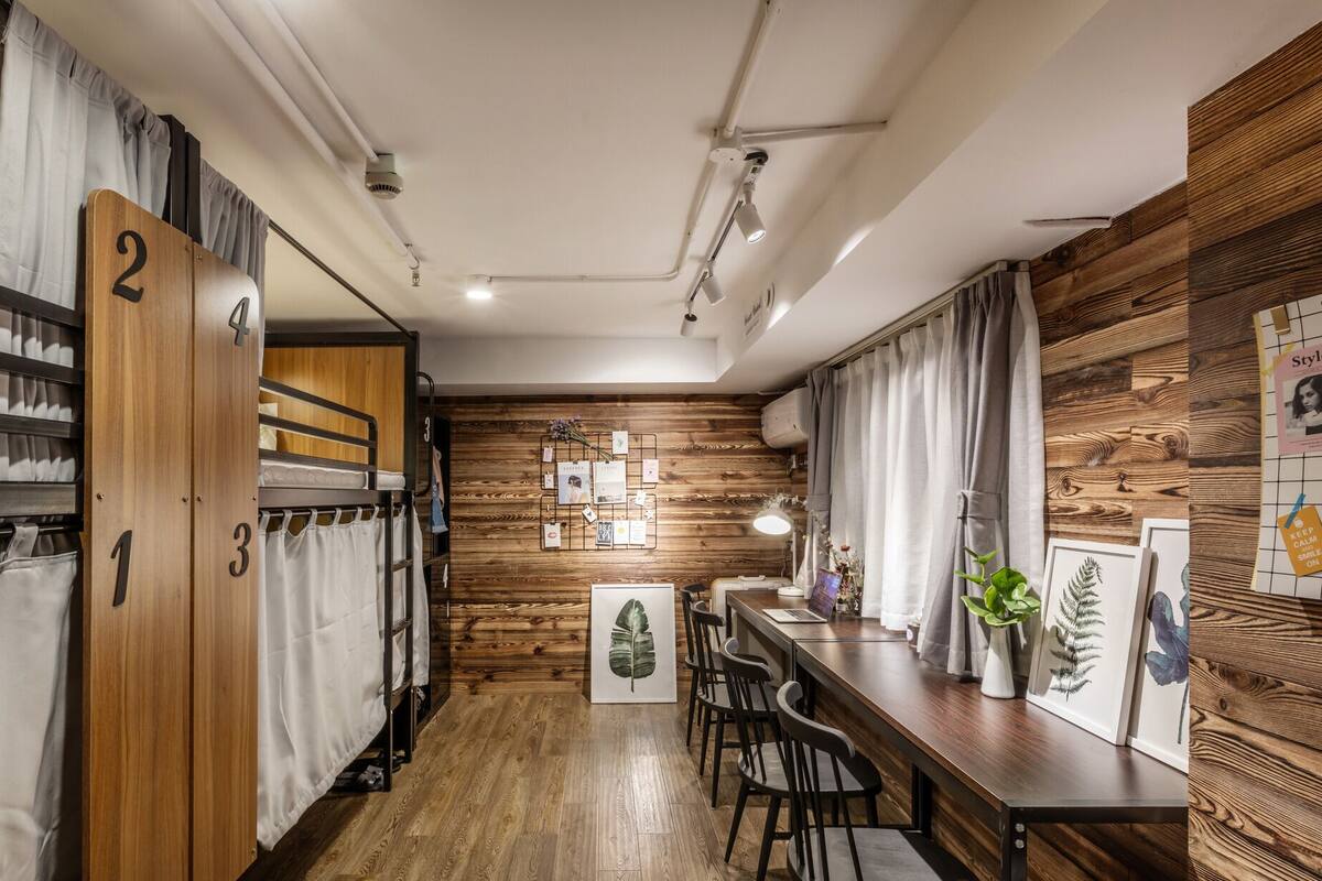 Una habitación de hotel compacta con literas, un panel de madera, un escritorio con sillas y un tablón de anuncios.