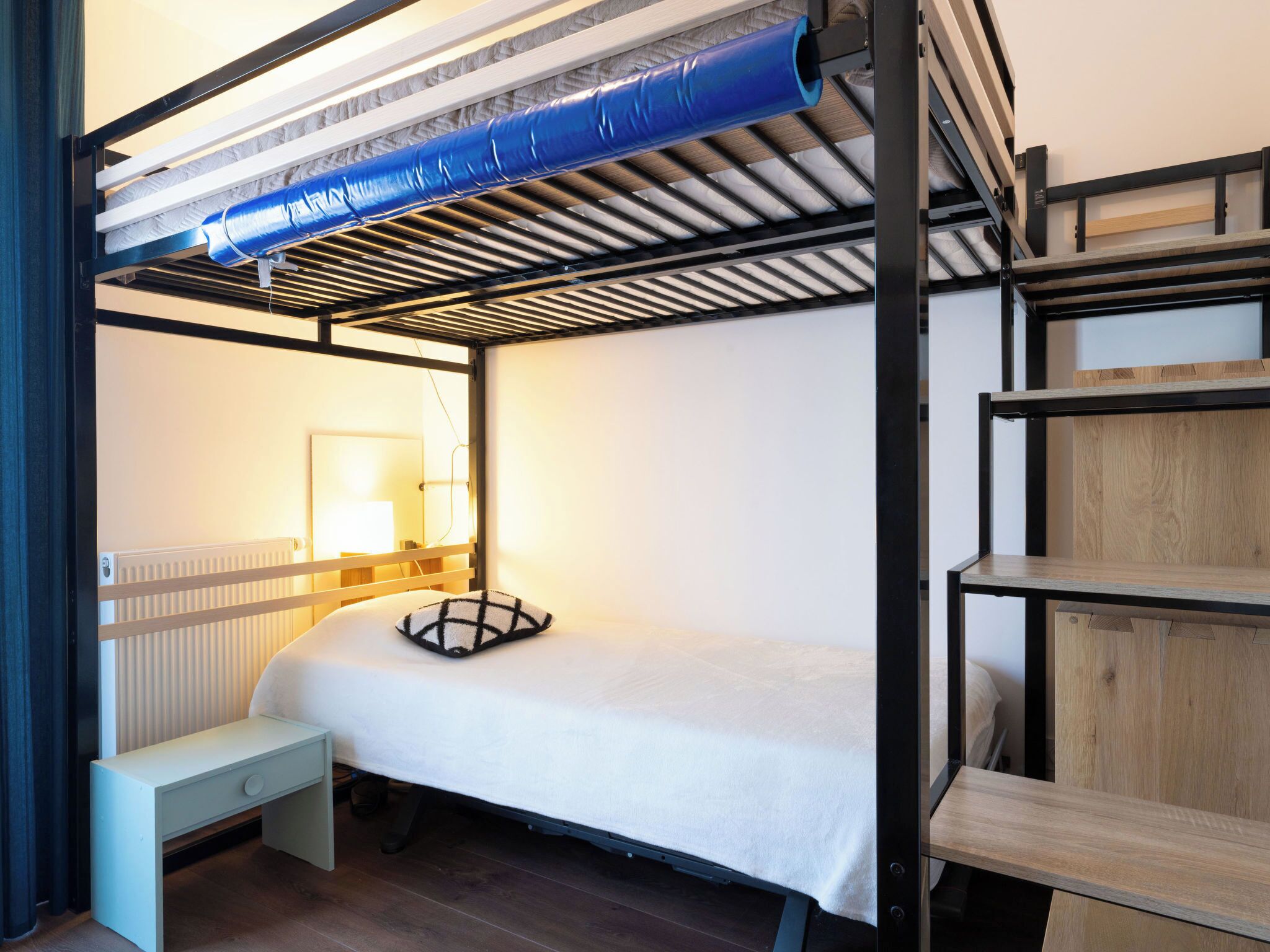 A compact, modern room with a bunk bed, a bedside table, a lamp, and a radiator.