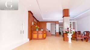 Un hall espacioso con un mostrador de recepción de madera, un jarrón blanco con flores y una zona de estar con sillas de madera.