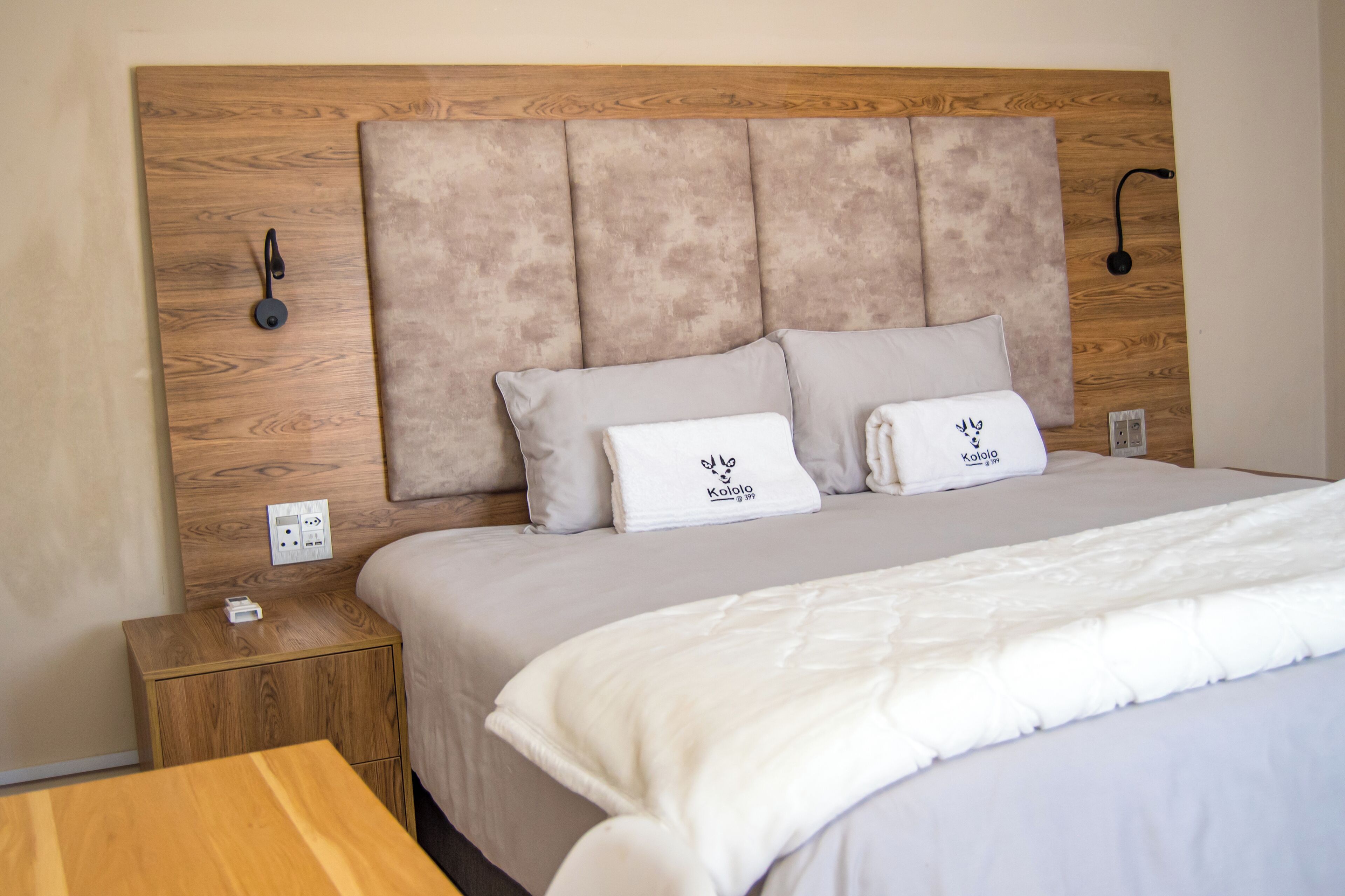 A neatly made bed with a wooden headboard, a bedside table with a power outlet, and a small wooden nightstand.