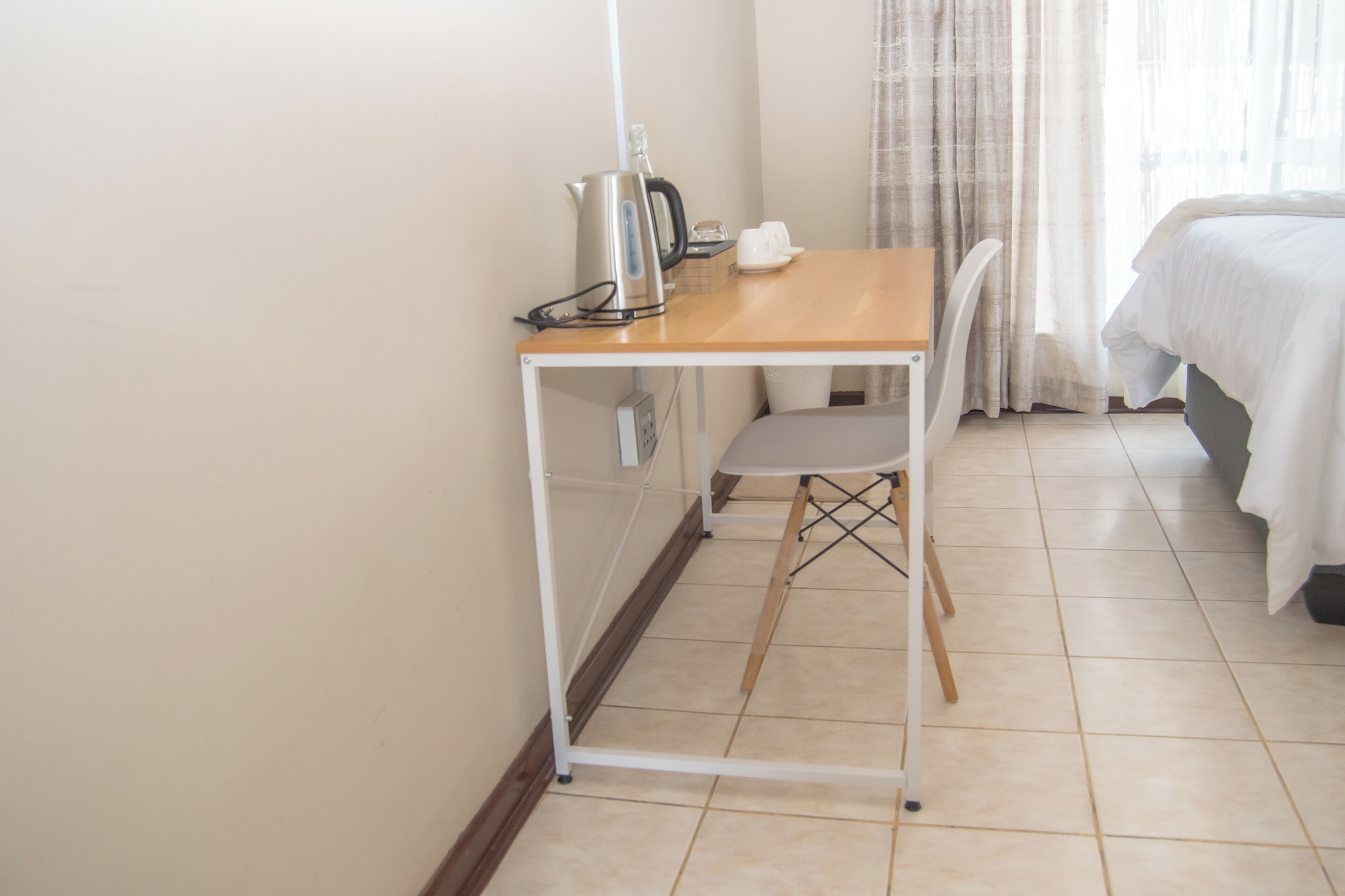 A small, clean room with a wooden desk, a chair, a bed, and a window with curtains.