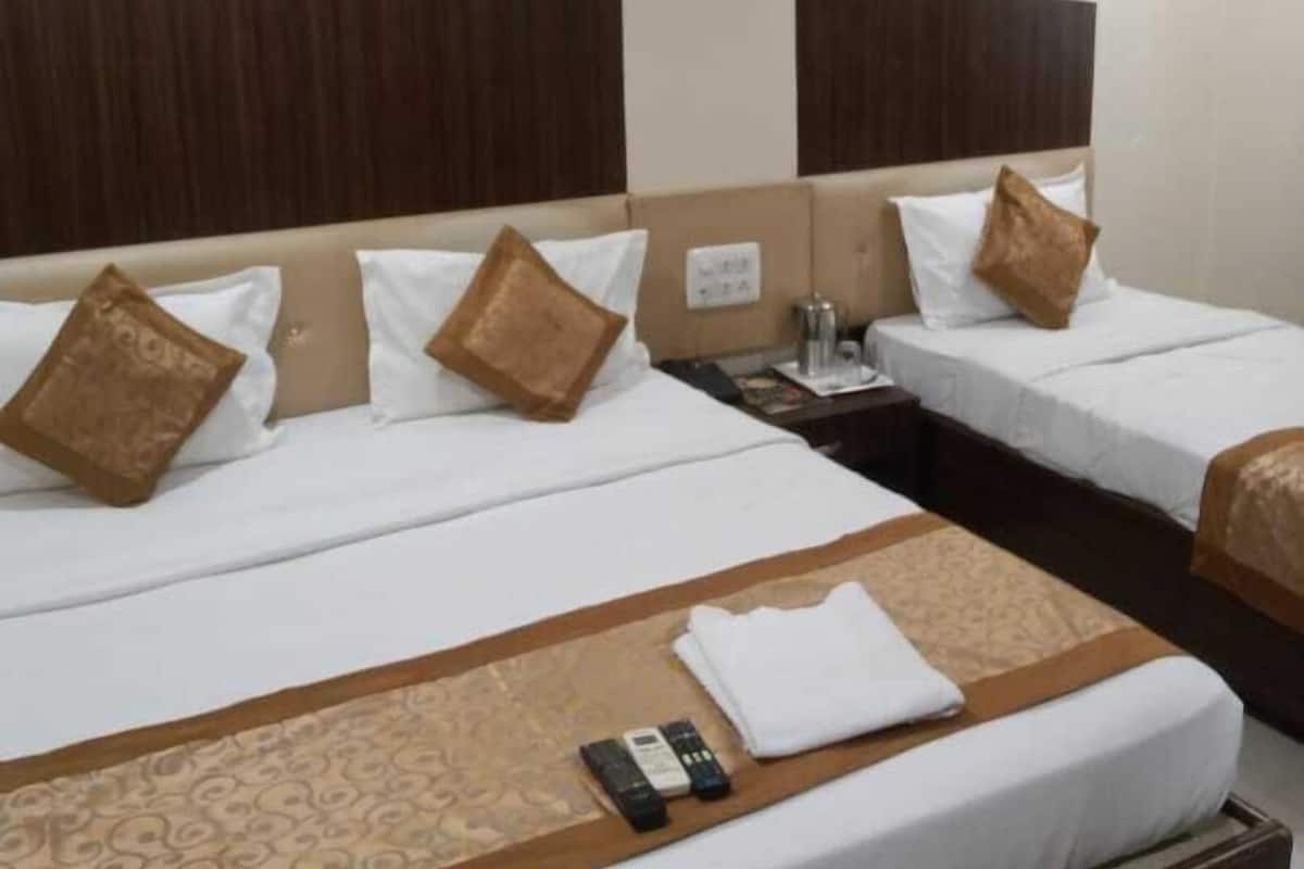 A hotel room with two beds, each with a wooden headboard, white bedding, and brown decorative pillows. There is a small table with a lamp, a telephone, and a remote control on the floor.