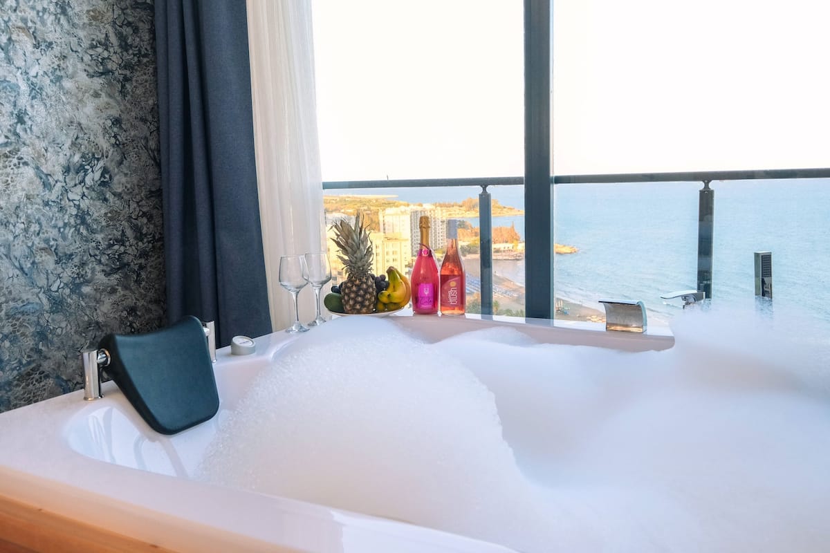 Une baignoire avec vue sur la mer, un panier de fruits et des bouteilles posées sur le rebord de la fenêtre.