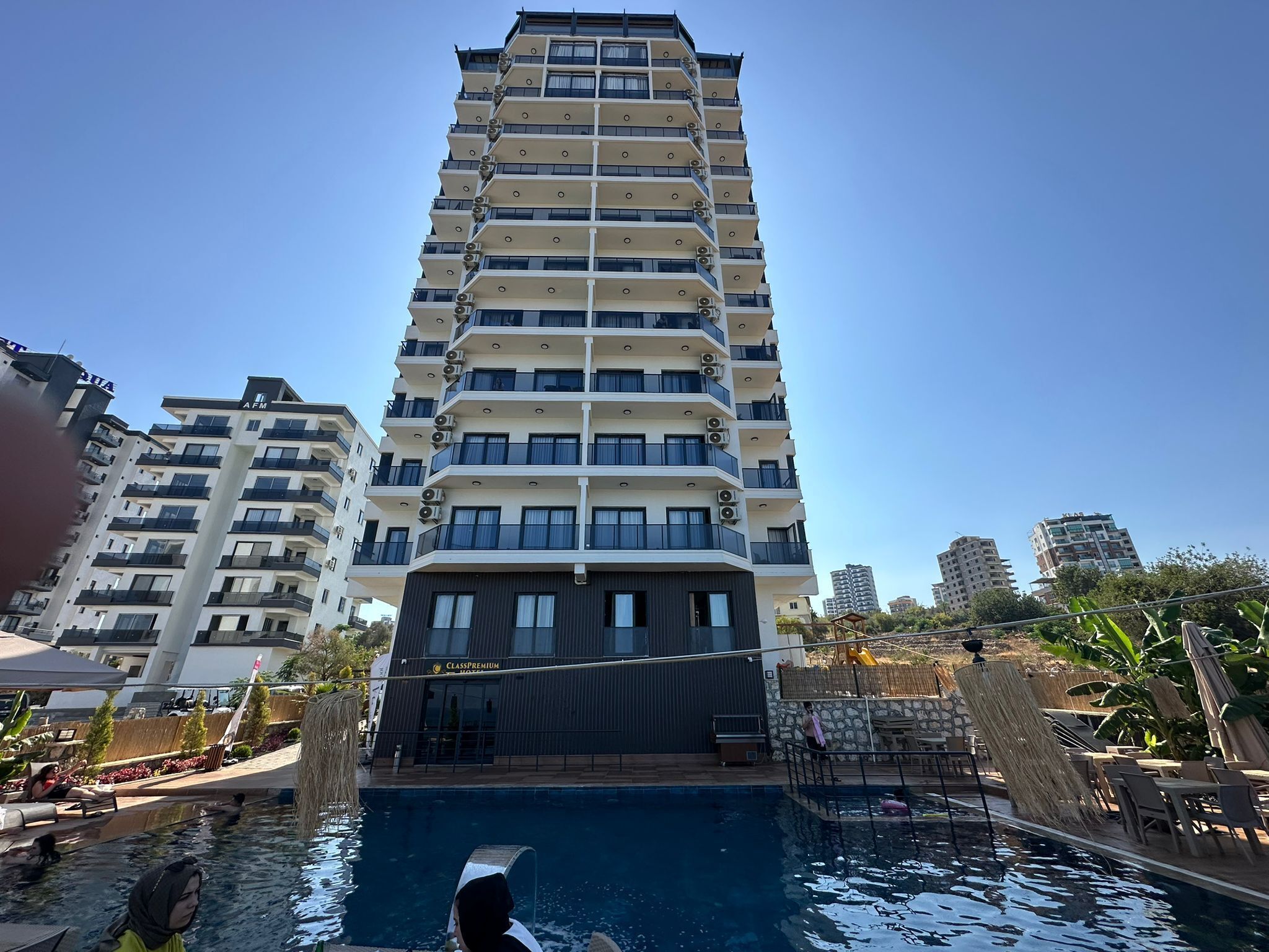 A high-rise building with multiple balconies and a swimming pool area with lounge chairs and umbrellas.