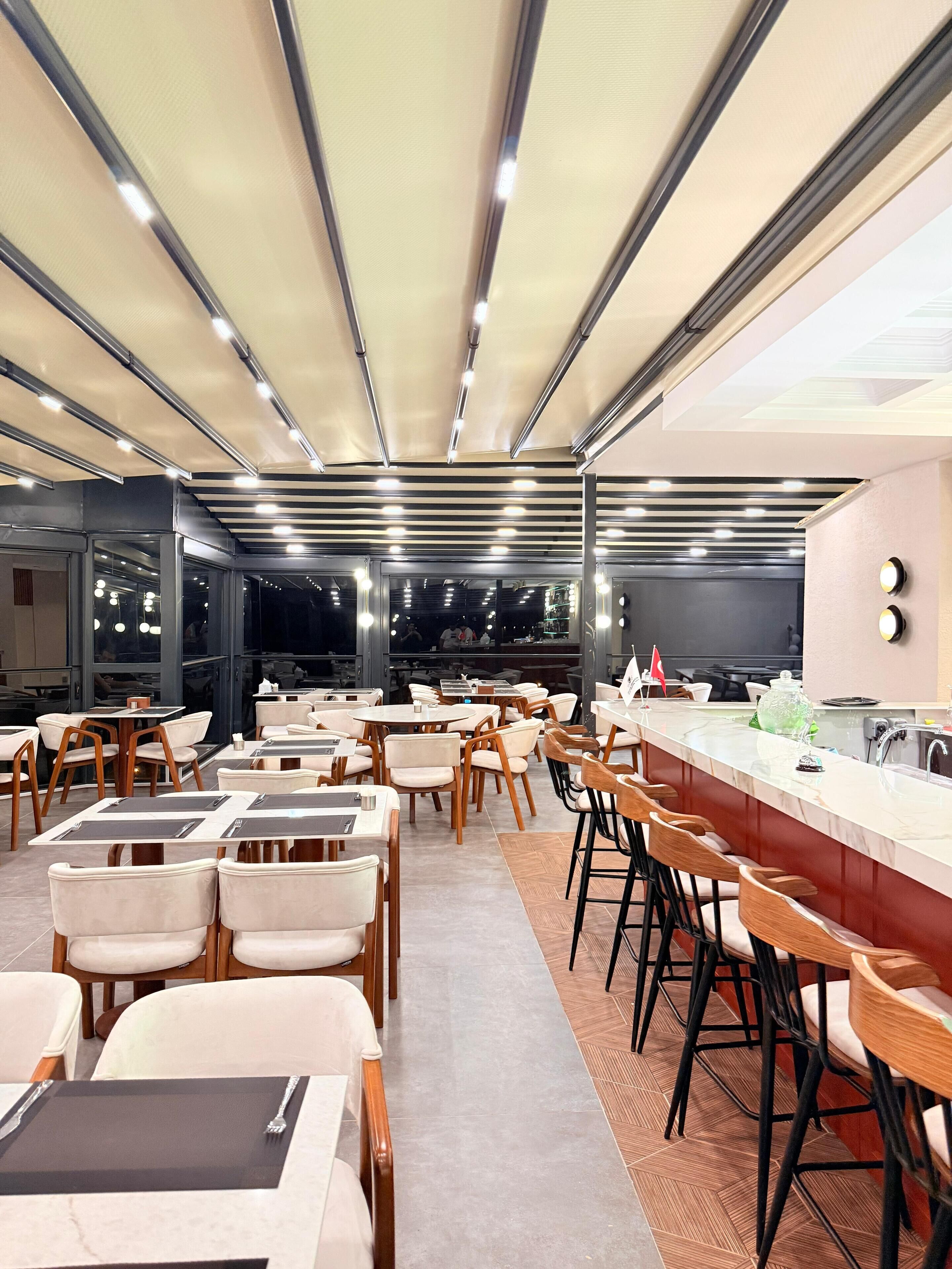 A modern restaurant with a long counter, multiple seating areas, and a high ceiling with strip lighting.