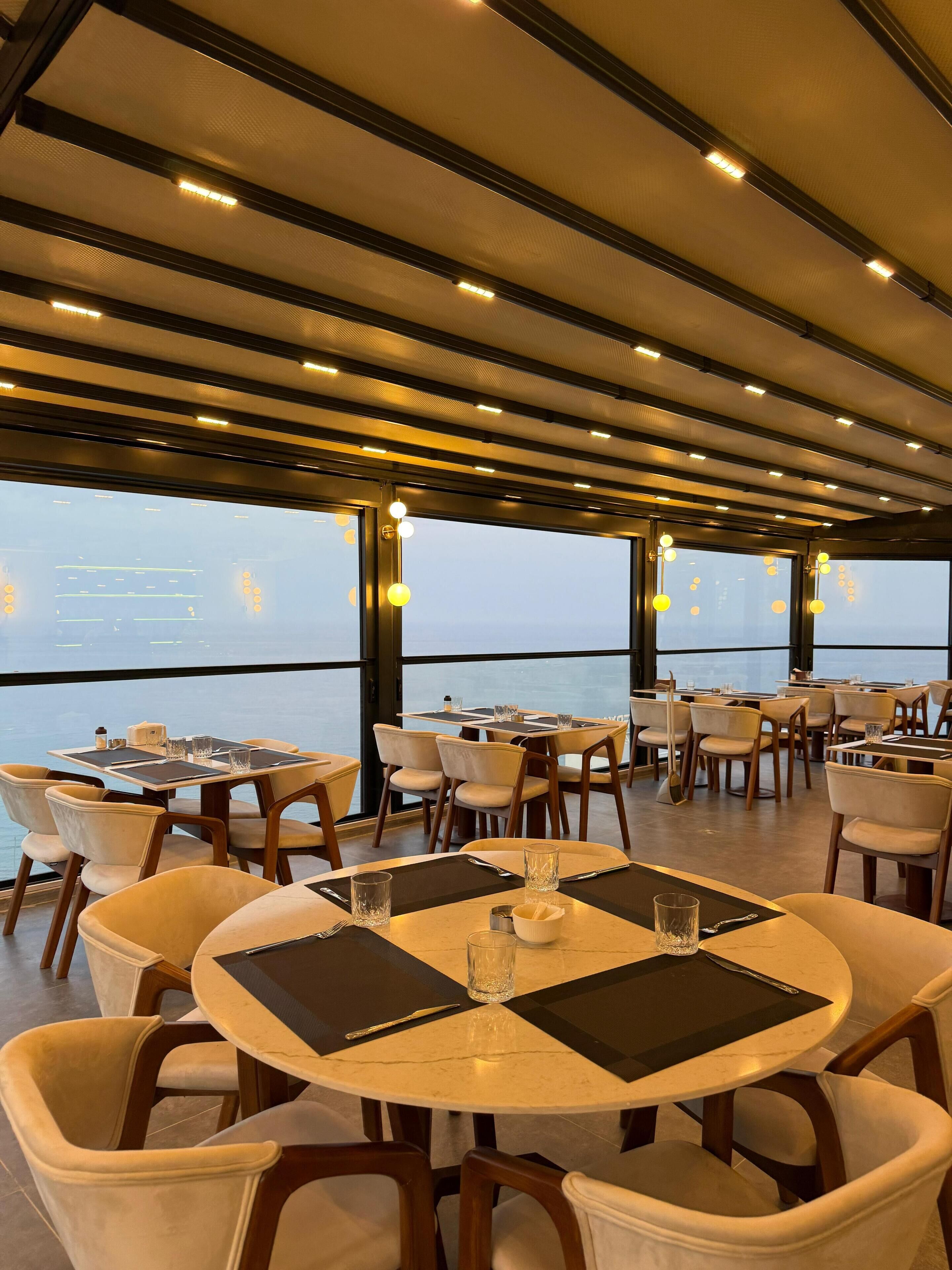 A restaurant with round tables and chairs, large windows, and a view of the sea.