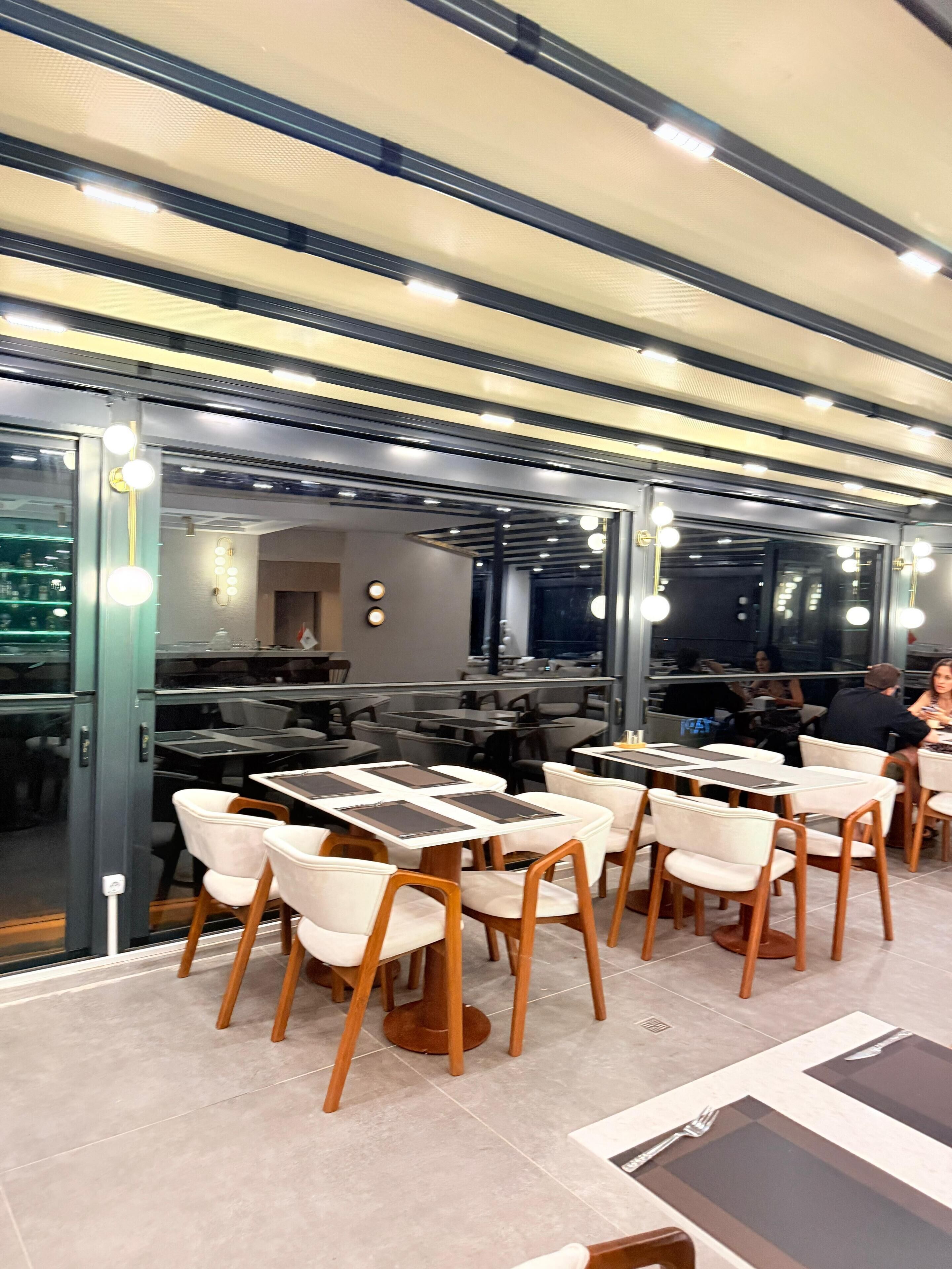 A restaurant with tables and chairs, a ceiling with lights, and a glass wall.