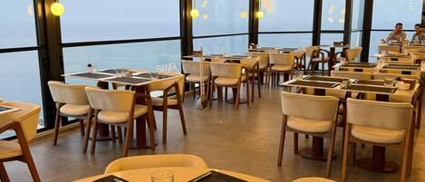 A restaurant with round tables, chairs, and a view of the sea.