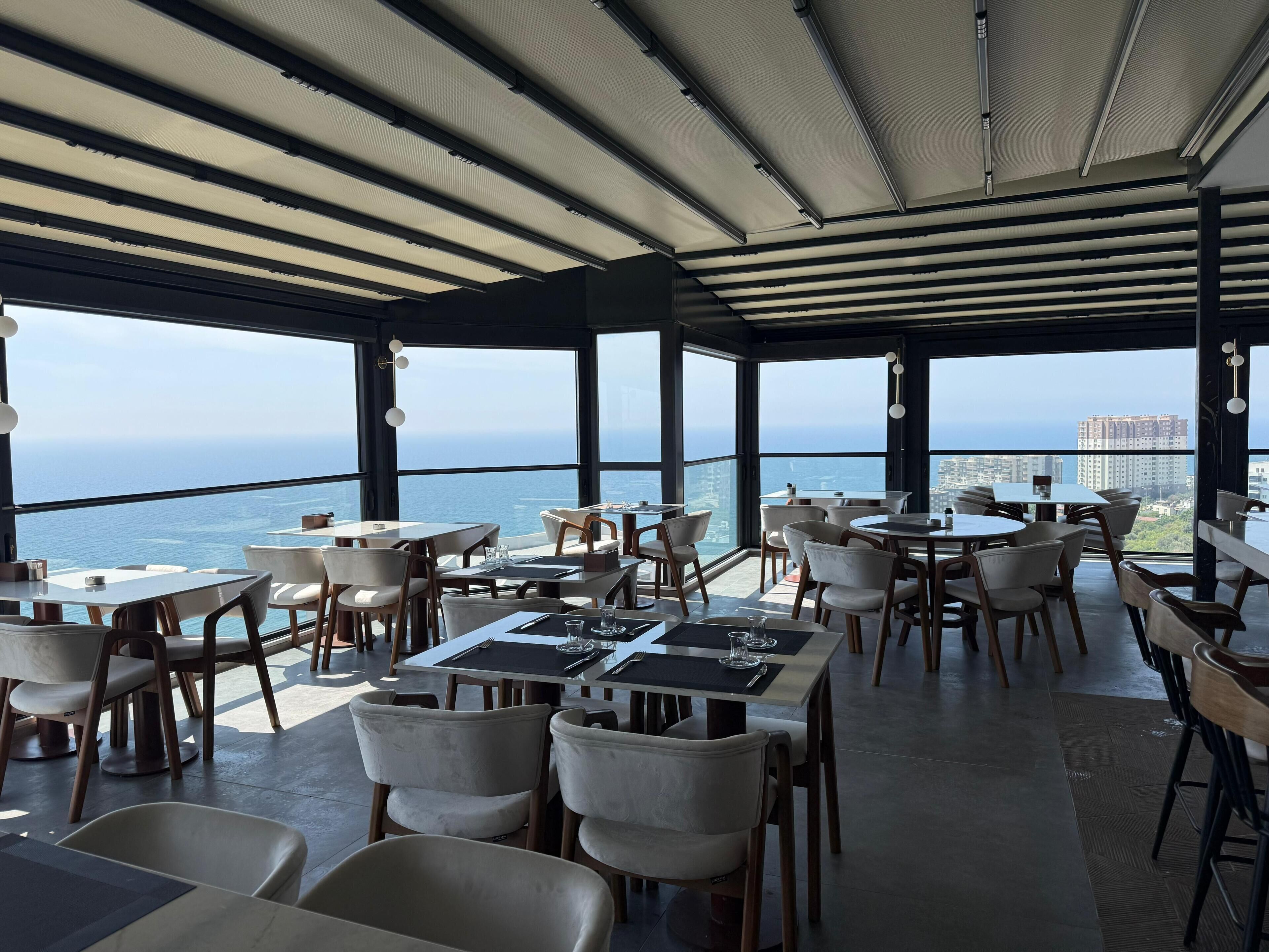 A restaurant with tables and chairs arranged under a large, open ceiling with a view of the ocean and cityscape.