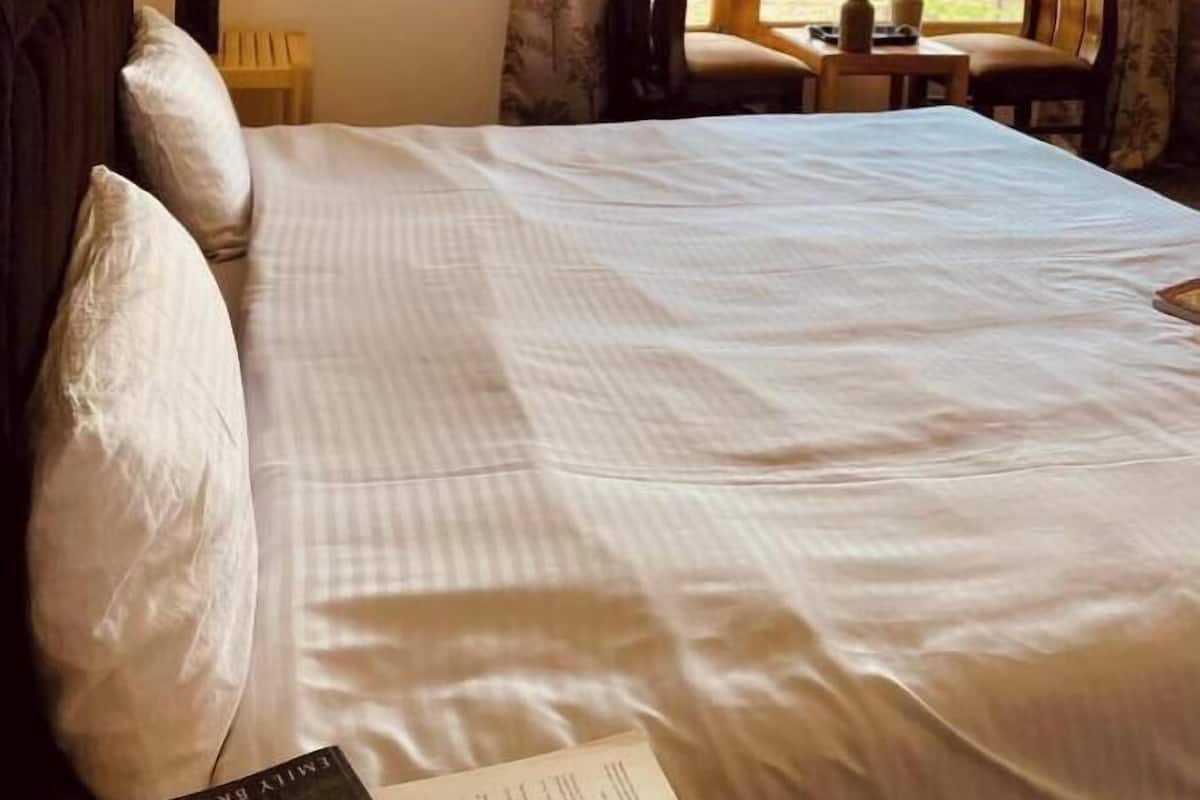 A neatly made bed with white linens, a wooden headboard, and a view of the outdoors through a window with curtains.