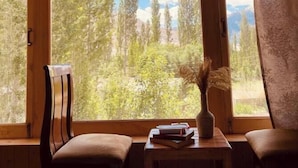 Habitación con un ventanal grande que ofrece vistas a árboles y montañas. Incluye una cama, dos sillas y una mesita con un florero y libros.