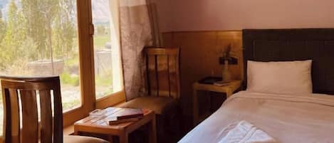 Ein gemütliches Schlafzimmer mit Holzbalkendecke, einem großen Bett, einem Sessel, einem kleinen Tisch und einem Fenster mit Blick auf Bäume und Berge.