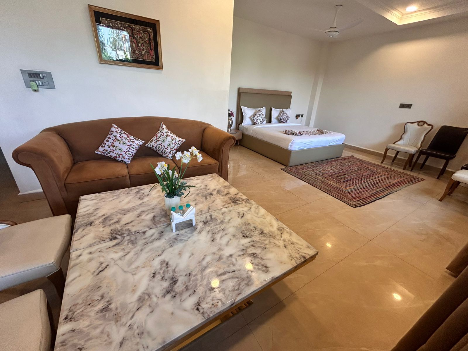 Una habitación de hotel moderna con una mesa de centro de mármol, un sofá marrón con cojines estampados, una cama con cabecera y una pequeña alfombra.