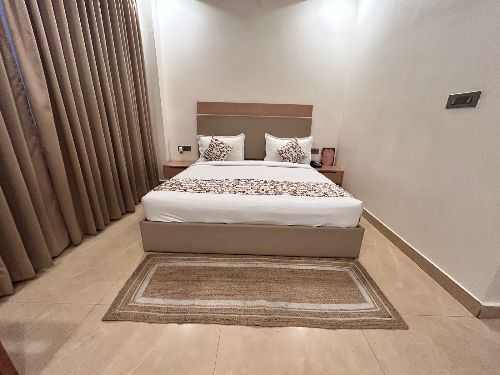 Una habitación de hotel con una cama grande, una mesita de noche, un teléfono en la pared y una ventana con cortinas.