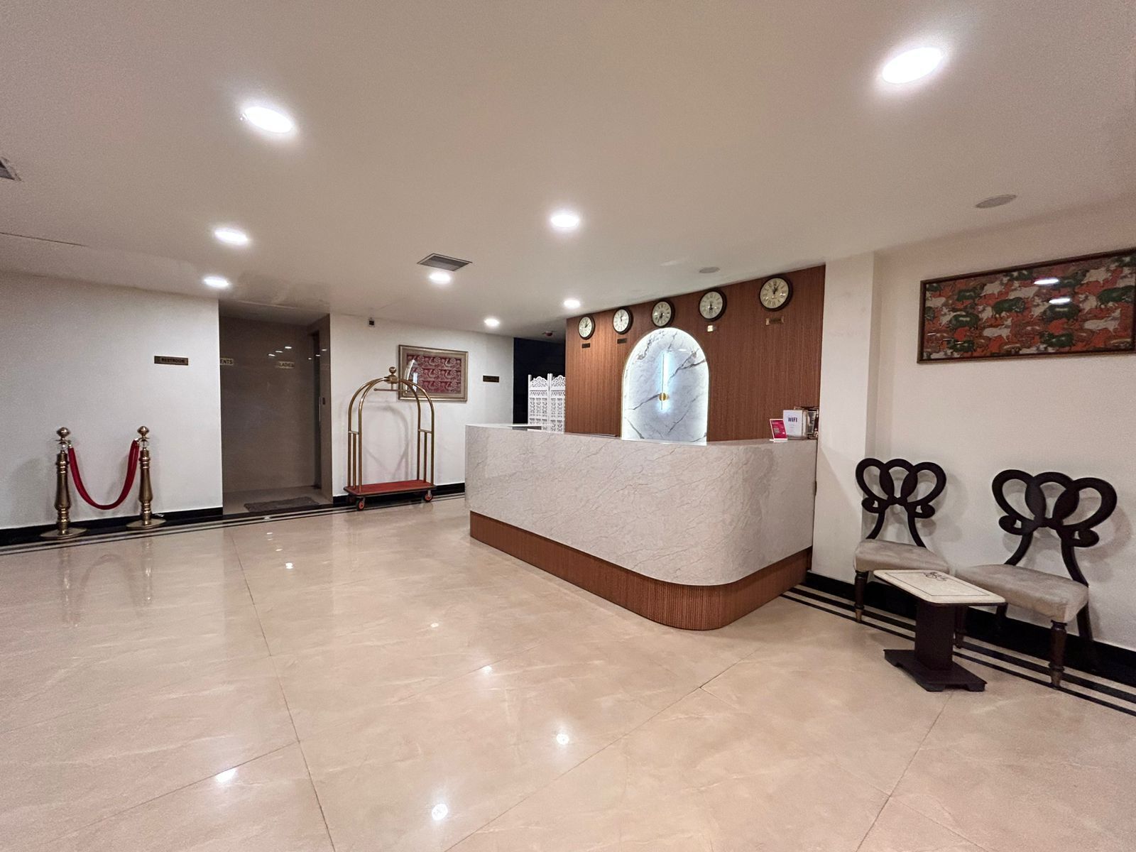 A hotel reception area with a marble counter, wall clocks, a reception desk, a red carpeted entrance, and a seating area with two chairs.
