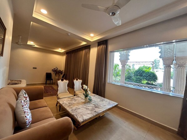 A living room with a sofa, a coffee table, a ceiling fan, and a window with a view of greenery. - Kairvi Fort Resort Moradabad (Moradabad)
