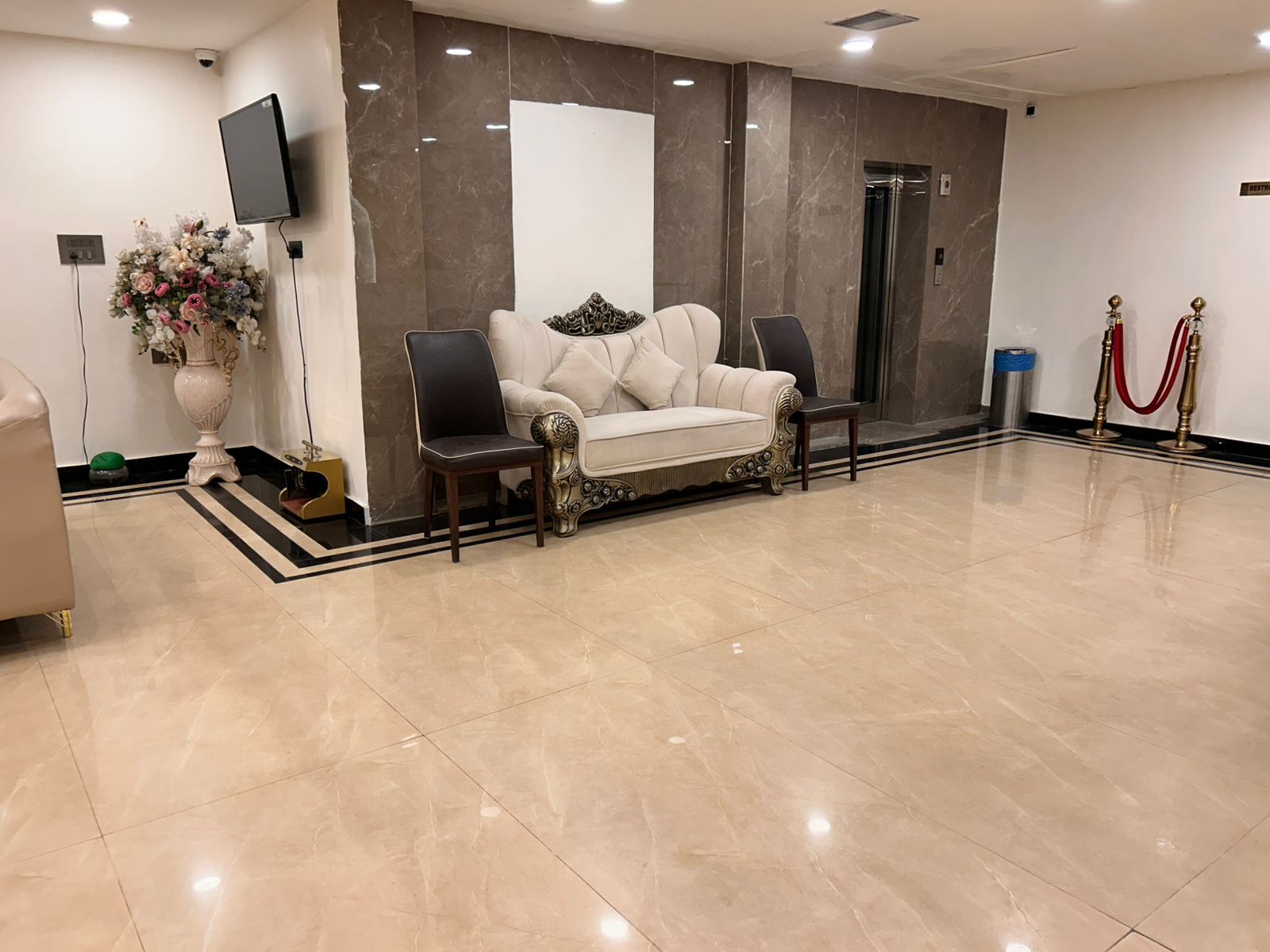 A lobby with a white sofa, a large vase of flowers, a flat-screen TV, a marble wall, and a red carpet.