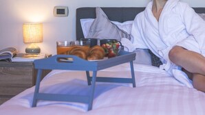 Une personne vêtue d’un peignoir blanc est assise sur un lit, avec un plateau contenant de la nourriture et des boissons.