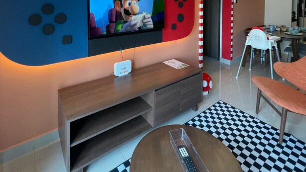 Ett modernt vardagsrum med en platt-TV som är monterad på en vägg med ett Mario-inspirerat motiv, en träinredd TV-möbel, ett rutmönstrat golv och en matplats med en röd, välvd öppning.