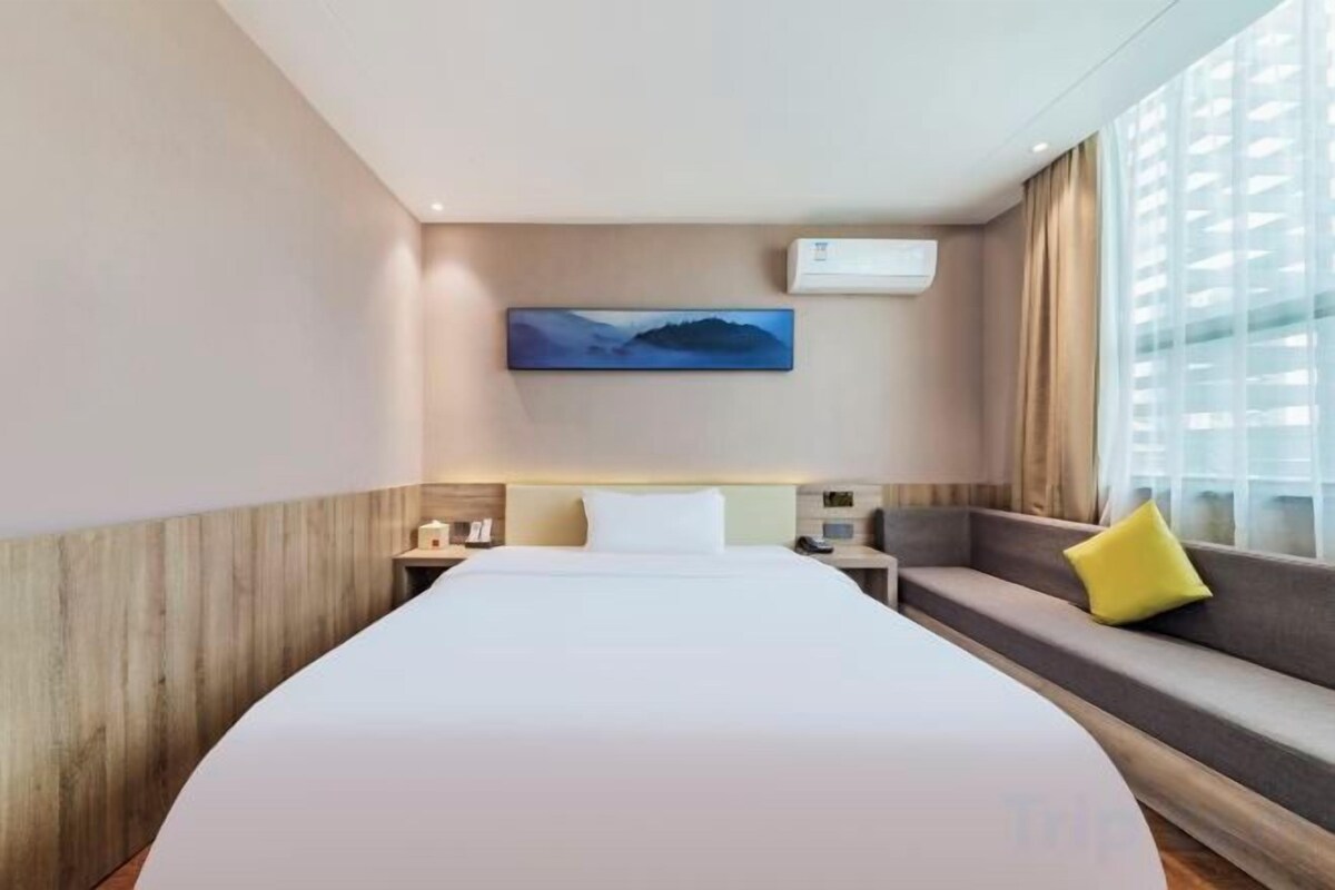 A hotel room with a large bed, a sofa with cushions, a painting on the wall, and a window with curtains.