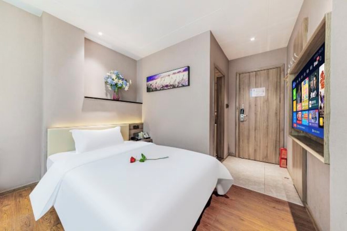 A hotel room with a large bed, a bedside table, a nightstand, a wall-mounted TV, and a wooden door.