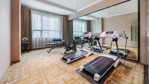 Une chambre d’hôtel dotée d’un grand miroir, d’équipements de fitness comprenant des tapis de course et des haltères, et offrant une vue sur la ville grâce à de grandes fenêtres.