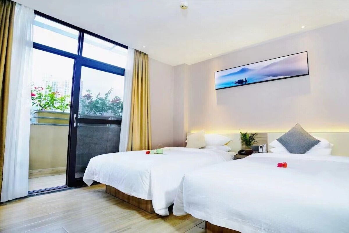 A hotel room with two beds, a sliding glass door to a balcony, a painting on the wall, and a nightstand with a lamp and a phone.