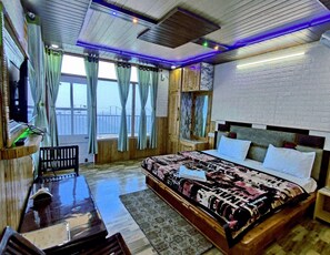A well-lit bedroom with a large bed, a desk, and a chair. There is a window with curtains and a view of the outside. - Hotel HILL Mount View Dalhousie Chamba  (Dalhousie)