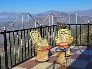 Two wicker chairs with colorful cushions on a balcony with a metal railing, overlooking a mountainous landscape. - Hotel HILL Mount View Dalhousie Chamba  (Dalhousie)